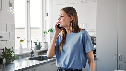 En ung tjej står i sitt kök och pratar i mobiltelefon. Hon har en blå t-shirt och långt hår.
