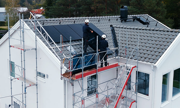 Bostadshus med byggställningar och två installatörer på taket som installerar solceller.