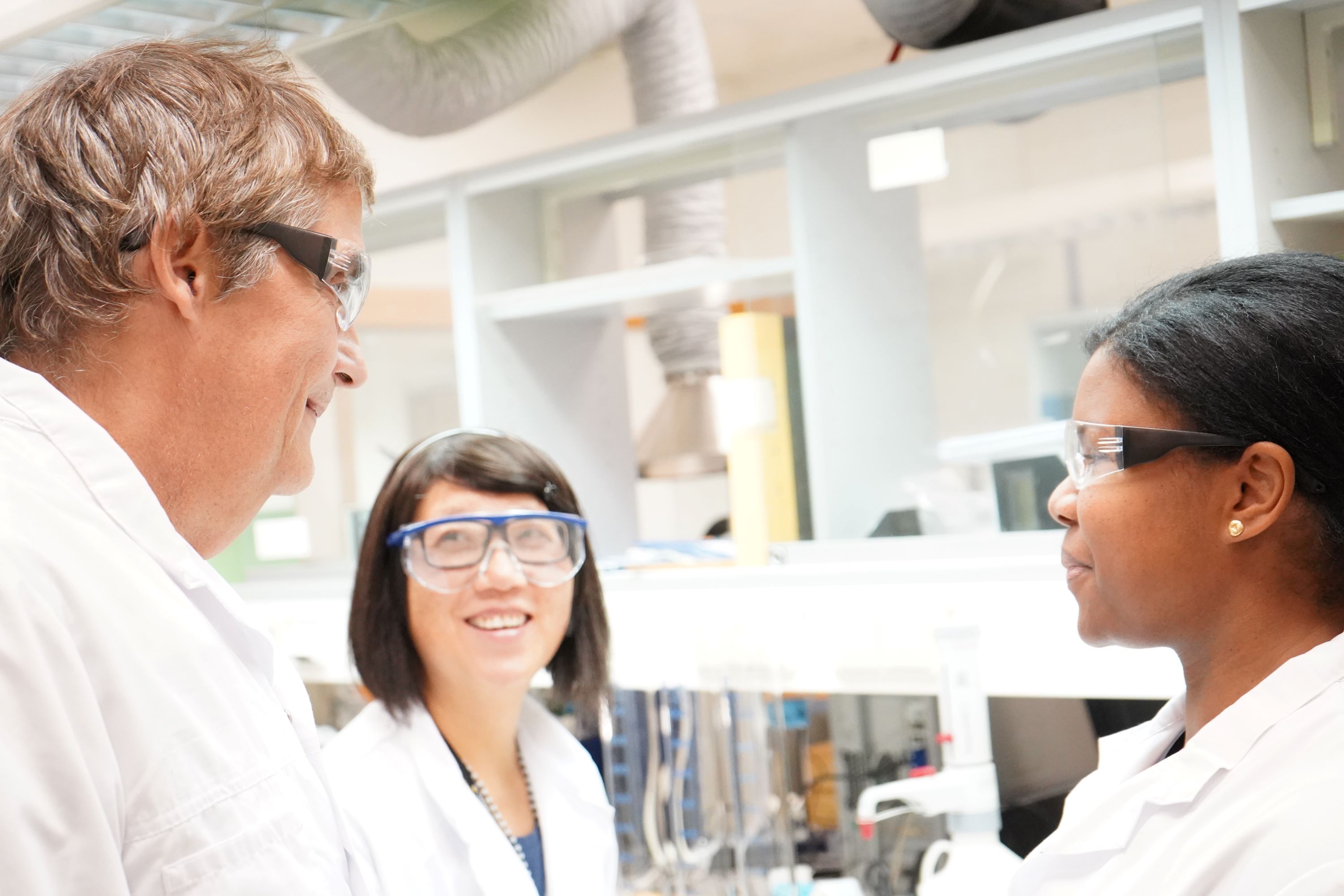 Tre personer med skyddsglasögon som står i ett laboratorium