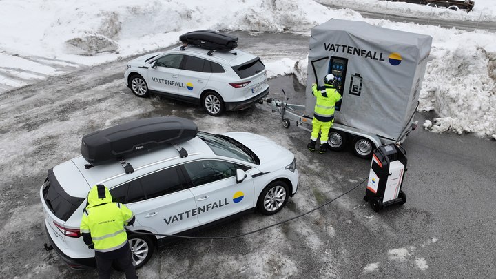 Två Vattenfall-bilar och en mobil energilagringstrailer på en snöig parkeringsplats, med två tekniker i varselkläder som arbetar med utrustningen.