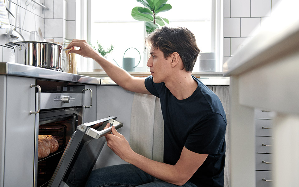 En man sitter på huk framför ugnen och kollar bröd medan en gryta kokar på spisen