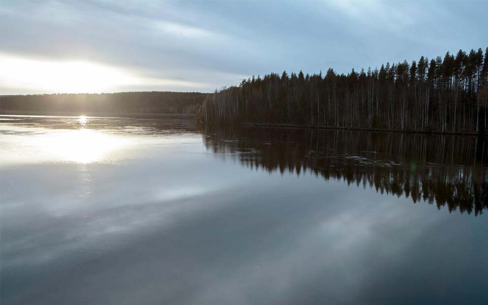 Solnedgång över stilla vatten med skog i bakgrunden och molnig himmel.