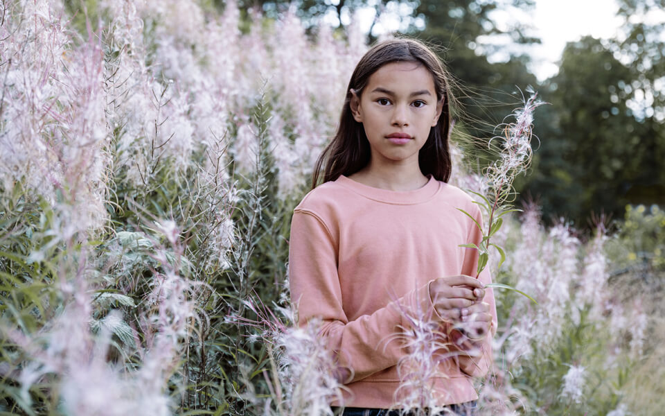 En ung tjej som står på en blommande äng. Hon har på sig en rosa tröja.