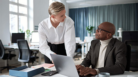 en man som sitter vid ett skrivbord och pratar med en kvinna som står bredvid honom