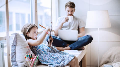 Far och dotter som sitter i en soffa. Flickan ritar på ett papper och pappan tittar i en laptop
