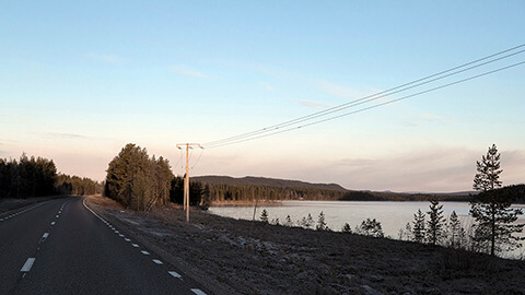 Landsväg intill en sjö, omgiven av skog och elledningar i mjukt kvälls- eller morgonljus.