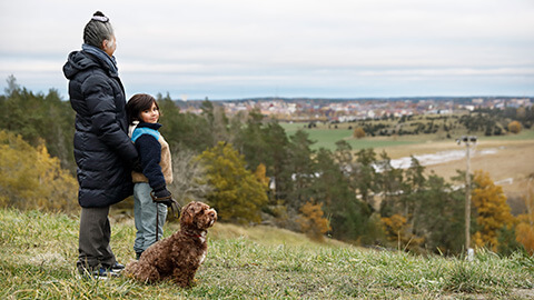 Kvinna, pojke och hund står på en kulle och tittar ut över ett höstlandskap med hus i bakgrunden
